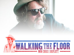 Bearded man wearing dark sunglasses and cowboy hat looks straight at camera.