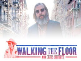 Bearded man with long hair wearing sunglasses against busy city street background.