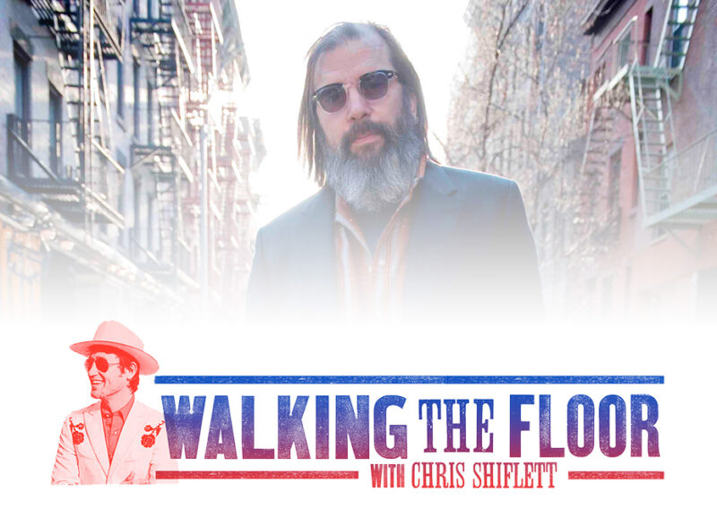 Bearded man with long hair wearing sunglasses against busy city street background.