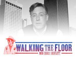 Man wearing glasses photographed before tall city buildings in black and white.