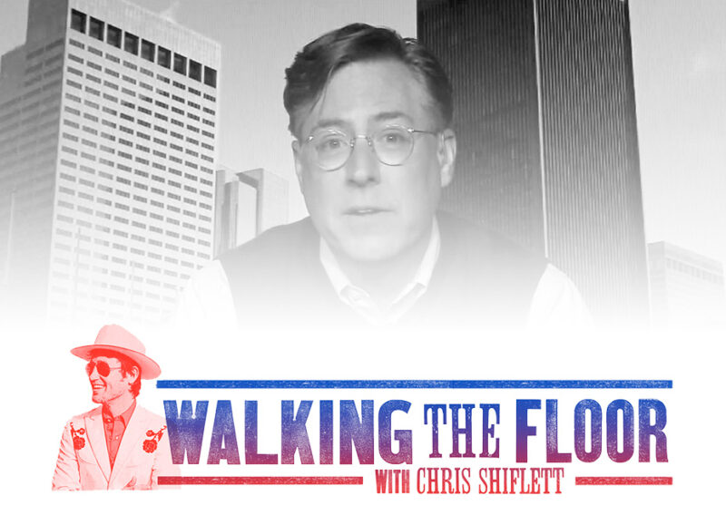 Man wearing glasses photographed before tall city buildings in black and white.