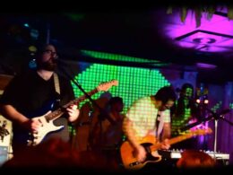 Musicians perform on a brightly lit stage with neon green walls and purple overhead lighting.