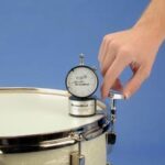 Hand holding a round analog drum tuner on a white drum head.