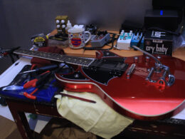 A red electric guitar sits on a workbench surrounded by guitar pedals and audio equipment.