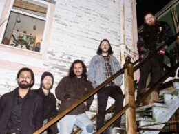 Five bearded men pose together on wooden stairs of a whitewashed building.