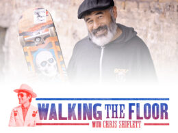 Man with gray beard in black cap and gray hoodie holding skateboard.
