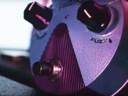 A Fuzz Face guitar effects pedal with control knobs is illuminated in purple and pink lighting.