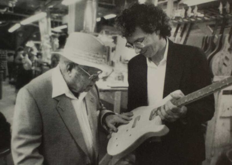 Black and white vintage photograph shows two men examining an electric guitar in a workshop setting with guitars in background.