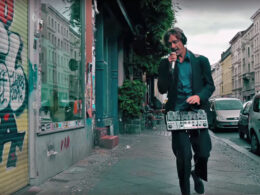 A man in a green jacket holds a looper pedal and microphone on a European street.