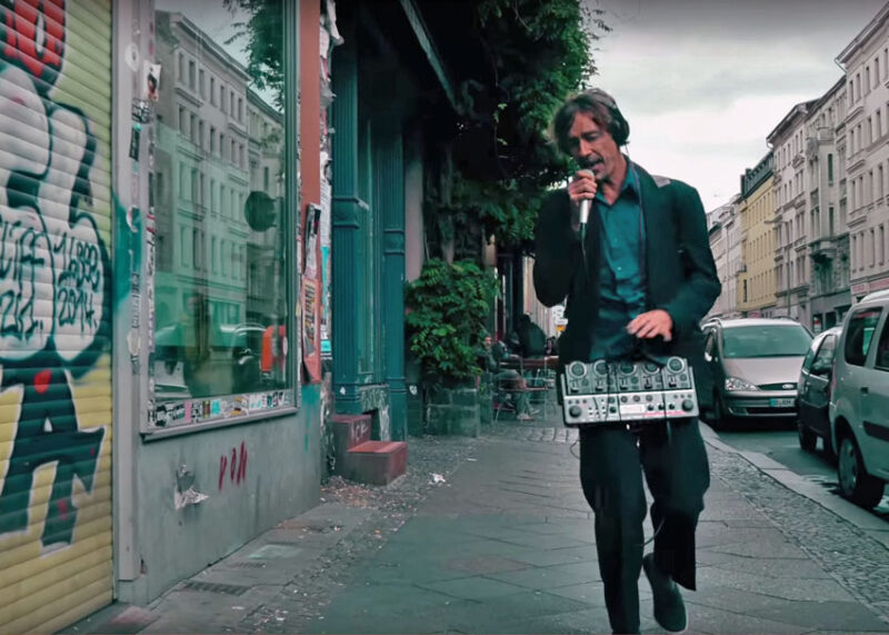 A man in a green jacket holds a looper pedal and microphone on a European street.