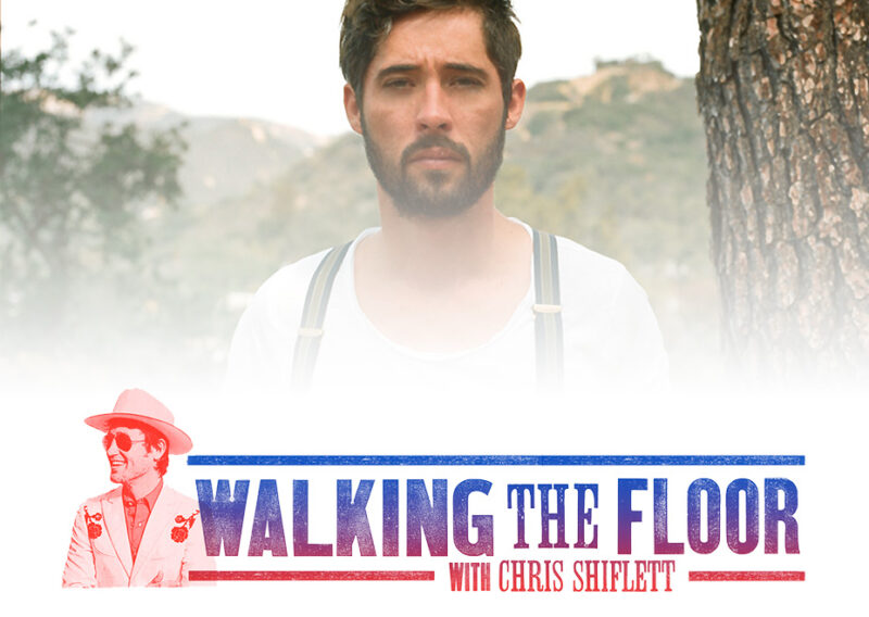 A man with a goatee wears a white tank top and shoulder straps outdoors.