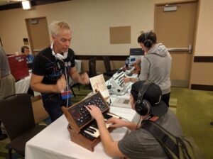 An instructor demonstrates a wooden synthesizer to a conference attendee.