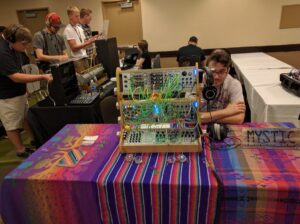 Multiple people gather around synthesizer equipment with colorful patch cables at an indoor event.