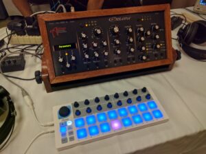 A wooden synthesizer with numerous controls sits above a white controller with illuminated blue buttons.