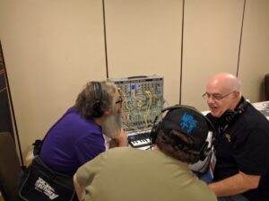 Three attendees listen to and discuss an analog modular synthesizer at an electronics gathering.