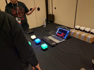 A presenter demonstrates glowing interactive devices and audio software at a table during an electronics exhibition.