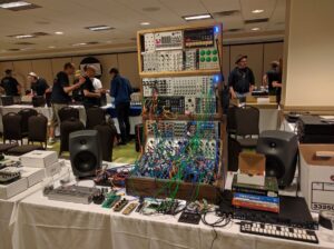 A large modular synthesizer installation features complex wiring, multiple keyboards, and speaker cabinets on wooden frames.