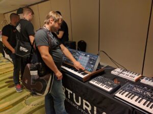 A man demonstrates synthesizer keyboards at a booth branded with a SYNTH logo and promotional materials.