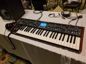 Deepmind 12 synthesizer with wood side panels on white table surrounded by cables.