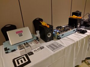 Electronic music vendor booth displaying laptops, synthesizers, and drum machines on white table.