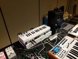 Music production desk features two white keyboard synthesizers, a black speaker, laptop, and various electronic equipment.