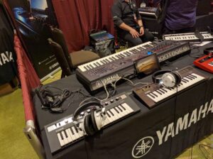 Yamaha synthesizers with colorful cables displayed on a black tablecloth at a convention.
