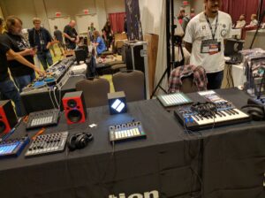 Multiple synthesizers and drum machines arranged on display tables at a music convention.
