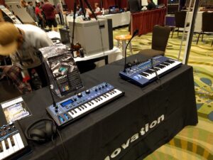 Two blue synthesizer keyboards displayed on a black tablecloth at a music equipment exhibition.