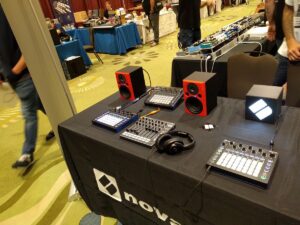 A Novation equipment booth displays audio controllers and red speakers on a convention floor table.