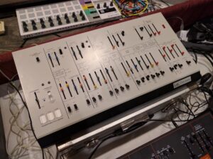 Close-up of a white modular synthesizer panel with sliders, knobs, and colorful patch cables.