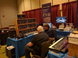 Two men demonstrate modular synthesizers at a booth with vintage audio equipment and signage.