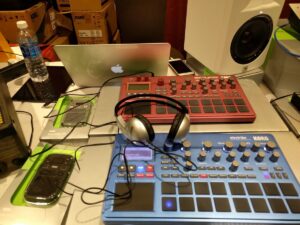 A music production desk features a MacBook, red and blue Korg synthesizers, and monitoring equipment.