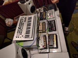 Synthesizers and drum modules with control knobs and cables are arranged on a white performance table.