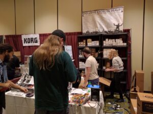 Music equipment vendors display synthesizers and products at a convention booth with exclusive promotional signage and deals.