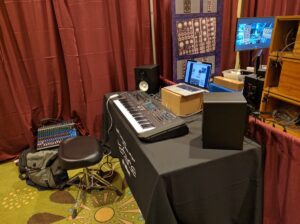 Music production studio featuring synthesizers, keyboards, mixing console, and displays with red curtains as acoustic treatment.