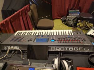 A Yamaha Montage synthesizer with mixer and headphones is set up behind red and black curtains.