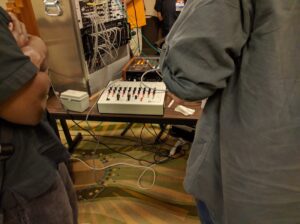 Professional audio synthesis equipment with controllers and cables is arranged with attendees at an event.