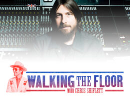 Bearded man sits at music recording console with many buttons visible.
