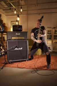 A tattooed guitarist plays a dark guitar in front of Marshall amplifiers.