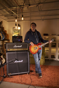 A musician holds a sunburst Les Paul guitar near Marshall amplifiers in a studio.
