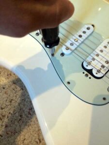 Hand using a screwdriver to remove a screw from a white guitar pickguard.