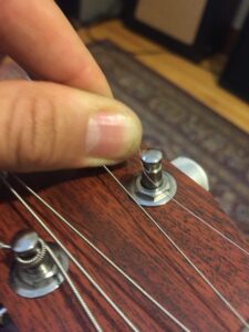 A finger touches an unwound plain string near a guitar tuning mechanism.