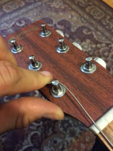 A hand touching a wound guitar string on a wooden headstock with tuning pegs.