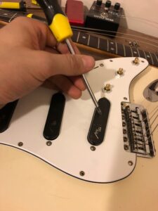 A person uses a yellow hammer to tap a potentiometer on a white electric guitar.