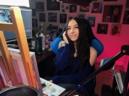 A woman in a blue top sits at a production desk in a pink-walled studio.