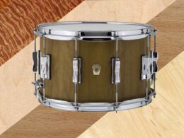 A snare drum with natural wood finish and chrome hardware is photographed against a textured background.