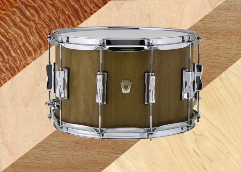 A snare drum with natural wood finish and chrome hardware is photographed against a textured background.