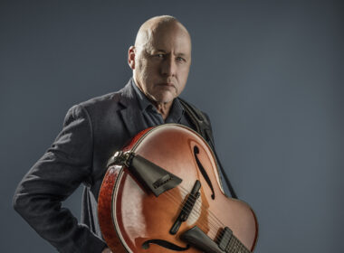 A musician holds a reddish-brown semi-acoustic guitar against a neutral gray background.