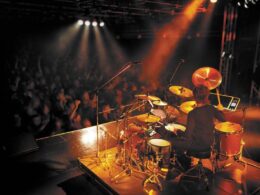 A drummer performing on a full drum kit under colored stage lighting.