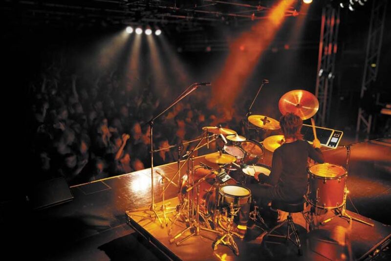 A drummer performing on a full drum kit under colored stage lighting.